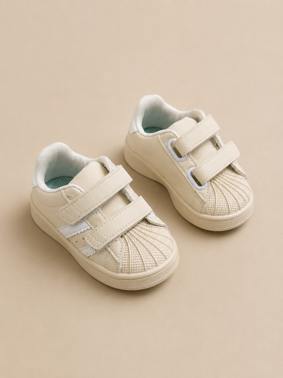 A pair of toddler's cream-colored velcro sneakers with a textured knit toe cap and light blue lining, set against a solid beige background.