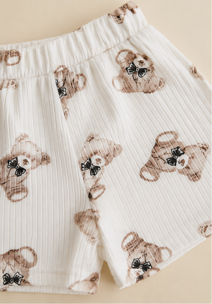A split view of a children's white ribbed knit outfit: a long-sleeve top with a large embroidered teddy bear patch on the chest, and matching shorts with a scattered teddy bear print. The background is split between white and a light beige tone.