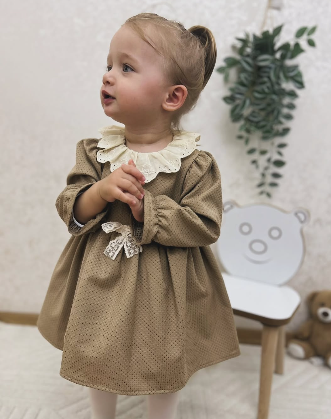 Child wearing a brown dress with a white collar in a room with a chair and plant.