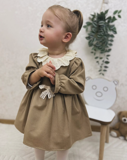 Child wearing a brown dress with a white collar in a room with a chair and plant.