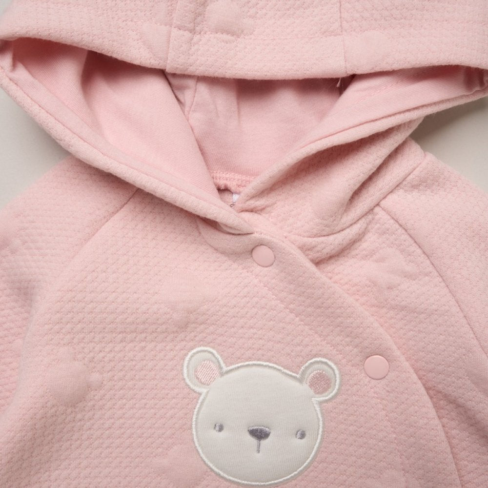 A close-up of a light pink hooded baby jacket or wrap with a textured waffle fabric, snap buttons, and a white embroidered teddy bear face on the front.