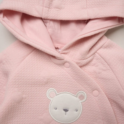 A close-up of a light pink hooded baby jacket or wrap with a textured waffle fabric, snap buttons, and a white embroidered teddy bear face on the front.