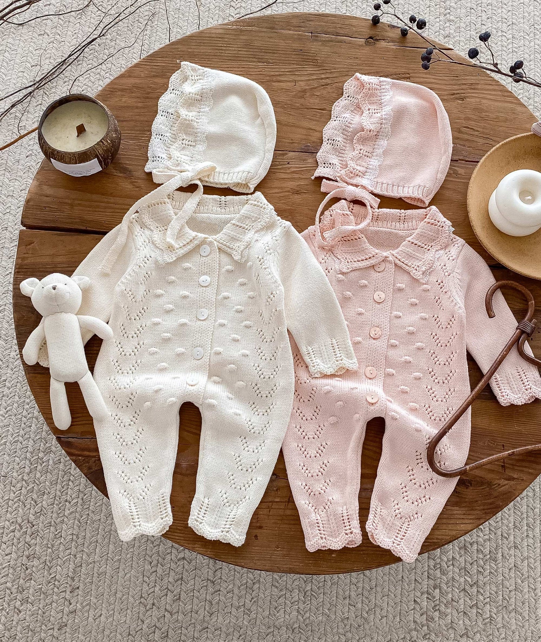 Two baby outfits, one white and one pink, on a wooden surface with matching hats and a teddy bear.