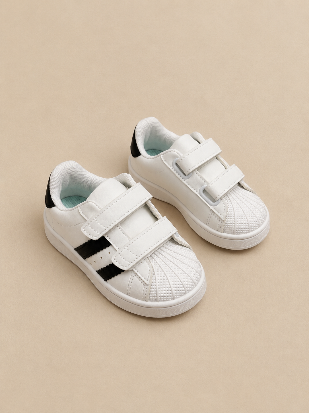 A pair of white children's sneakers with three hook-and-loop straps and black side stripes, displayed on a beige background.