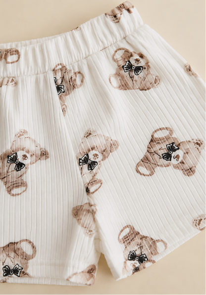A split view of a children's white ribbed knit outfit: a long-sleeve top with a large embroidered teddy bear patch on the chest, and matching shorts with a scattered teddy bear print. The background is split between white and a light beige tone.