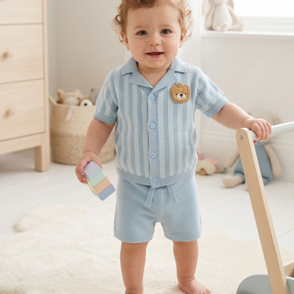 Baby in light blue striped outfit with bear emblem standing in a room with toys and furniture.